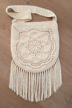 Cream crochet shoulder bag with a floral mandala pattern and fringe, laid flat on a light brown wooden floor.