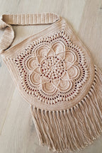 A beige crocheted shoulder bag with a large floral mandala pattern and long fringe, laid flat on a light wood floor.