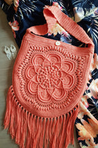 A handmade coral pink crochet shoulder bag with a large floral mandala pattern and long fringe, laid flat on a dark floral fabric background.