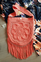 A coral orange crochet shoulder bag with a floral mandala design and long fringe, laid flat on a wooden surface next to a navy floral blanket.
