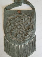 A handmade crocheted shoulder bag in a muted sage green color, featuring an intricate floral mandala pattern on the front, a single wooden button closure, and long fringe at the bottom, displayed on a white wooden plank background.