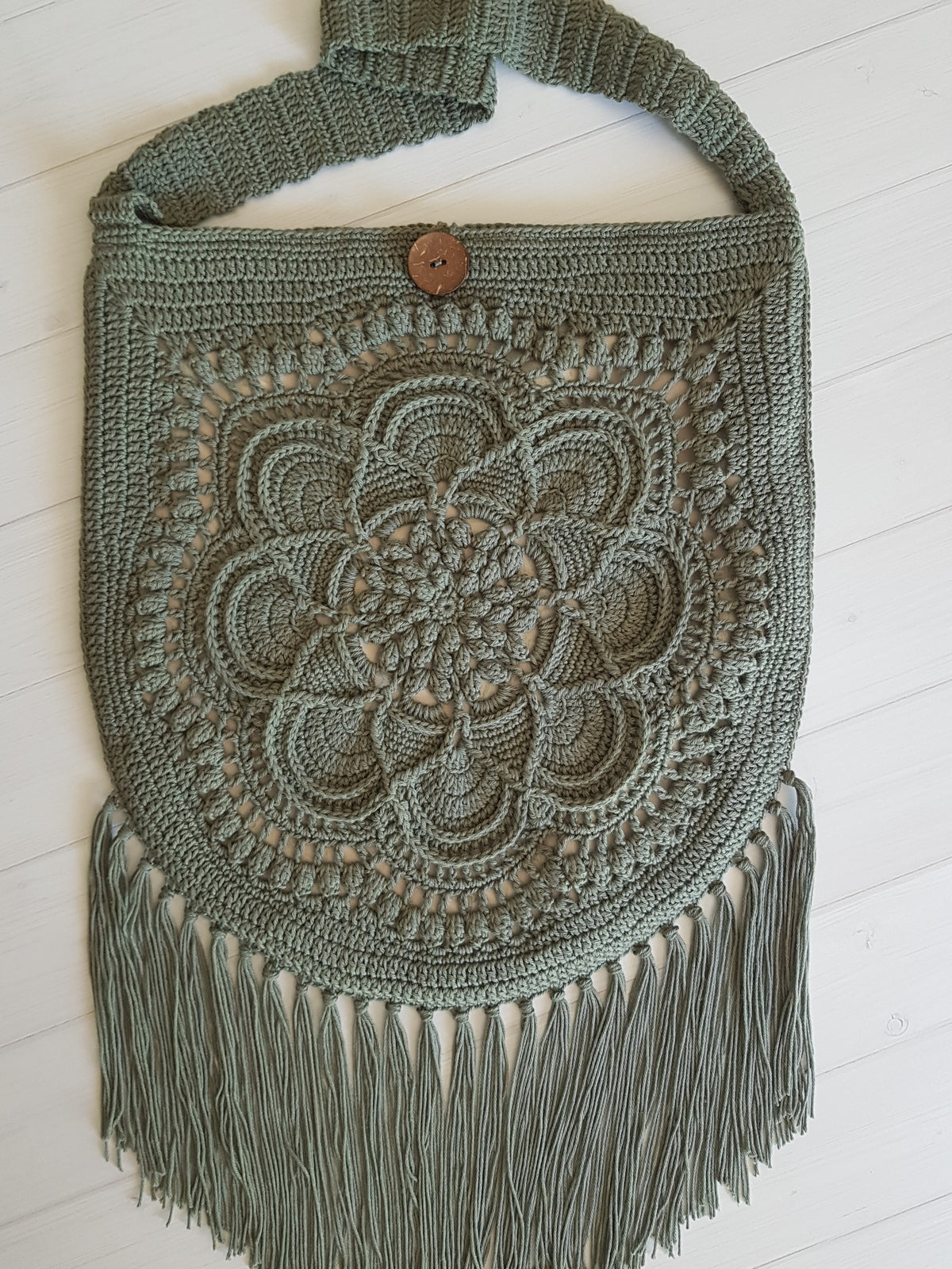 A handmade crocheted shoulder bag in a muted sage green color, featuring an intricate floral mandala pattern on the front, a single wooden button closure, and long fringe at the bottom, displayed on a white wooden plank background.