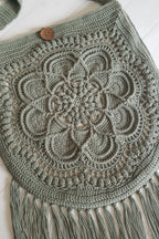 A sage green crocheted shoulder bag with a large floral mandala pattern, fringe tassels along the bottom, and a single wooden button closure, laid flat on a white wood background.