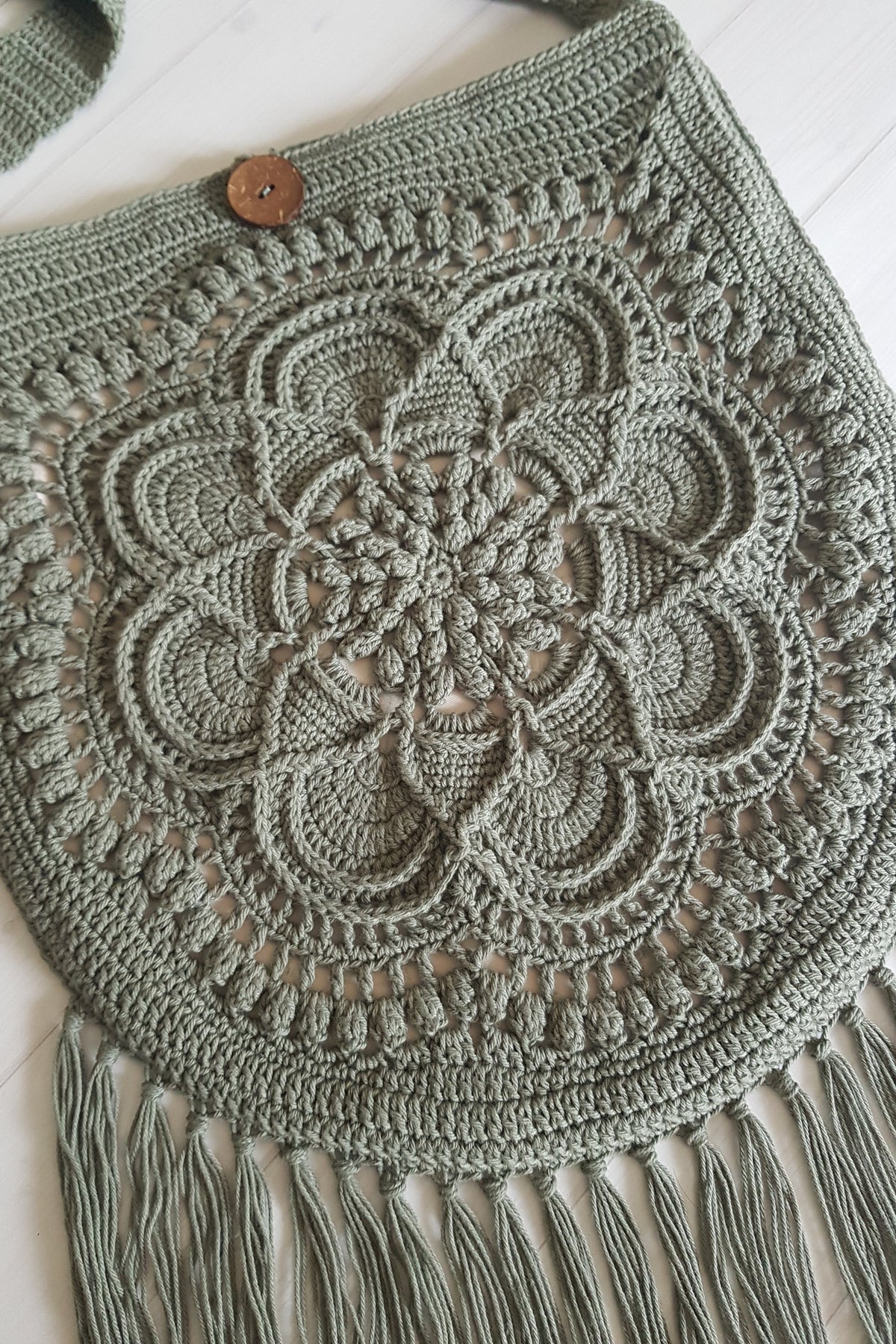 A sage green crocheted shoulder bag with a large floral mandala pattern, fringe tassels along the bottom, and a single wooden button closure, laid flat on a white wood background.