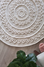 A round, off-white crocheted rug with intricate concentric mandala patterns, laid out on a light wood floor with a green plant and a pink cushion visible in the foreground.