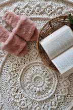 A round cream-colored crocheted rug with intricate lace and floral patterns, styled with a pair of dusty pink slippers and a wicker basket holding an open book on a light wood floor.