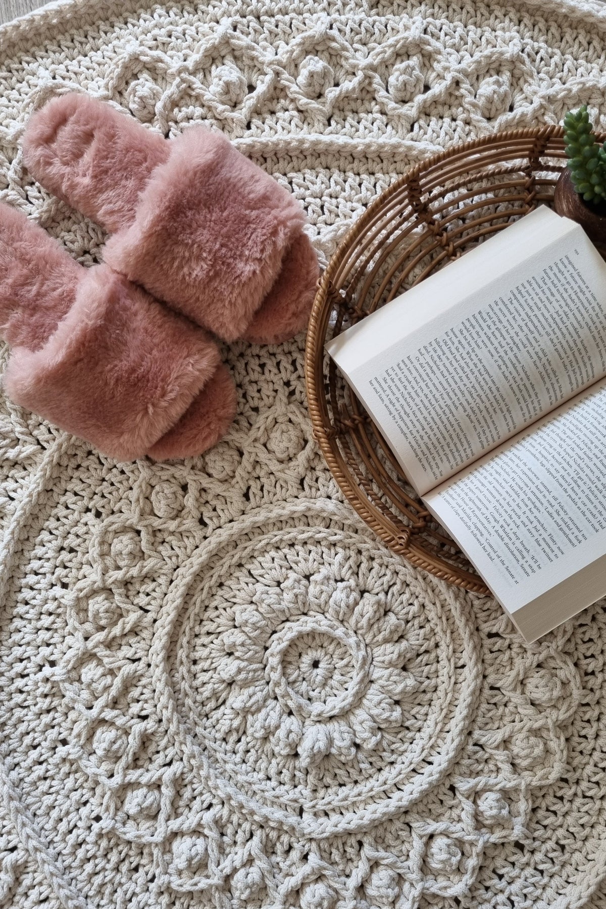 A round cream-colored crocheted rug with intricate lace and floral patterns, styled with a pair of dusty pink slippers and a wicker basket holding an open book on a light wood floor.