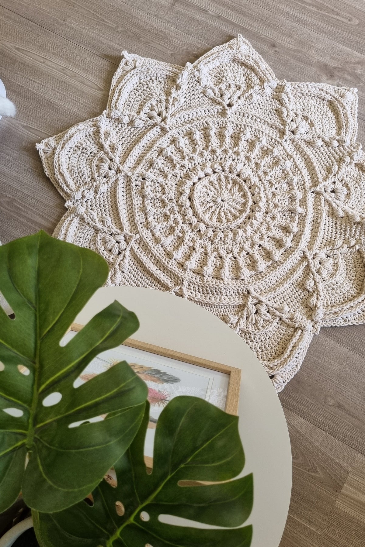 A round, off-white crocheted doily with intricate mandala-like patterns, styled on a light wood floor next to a white table with a green monstera plant and a picture frame.