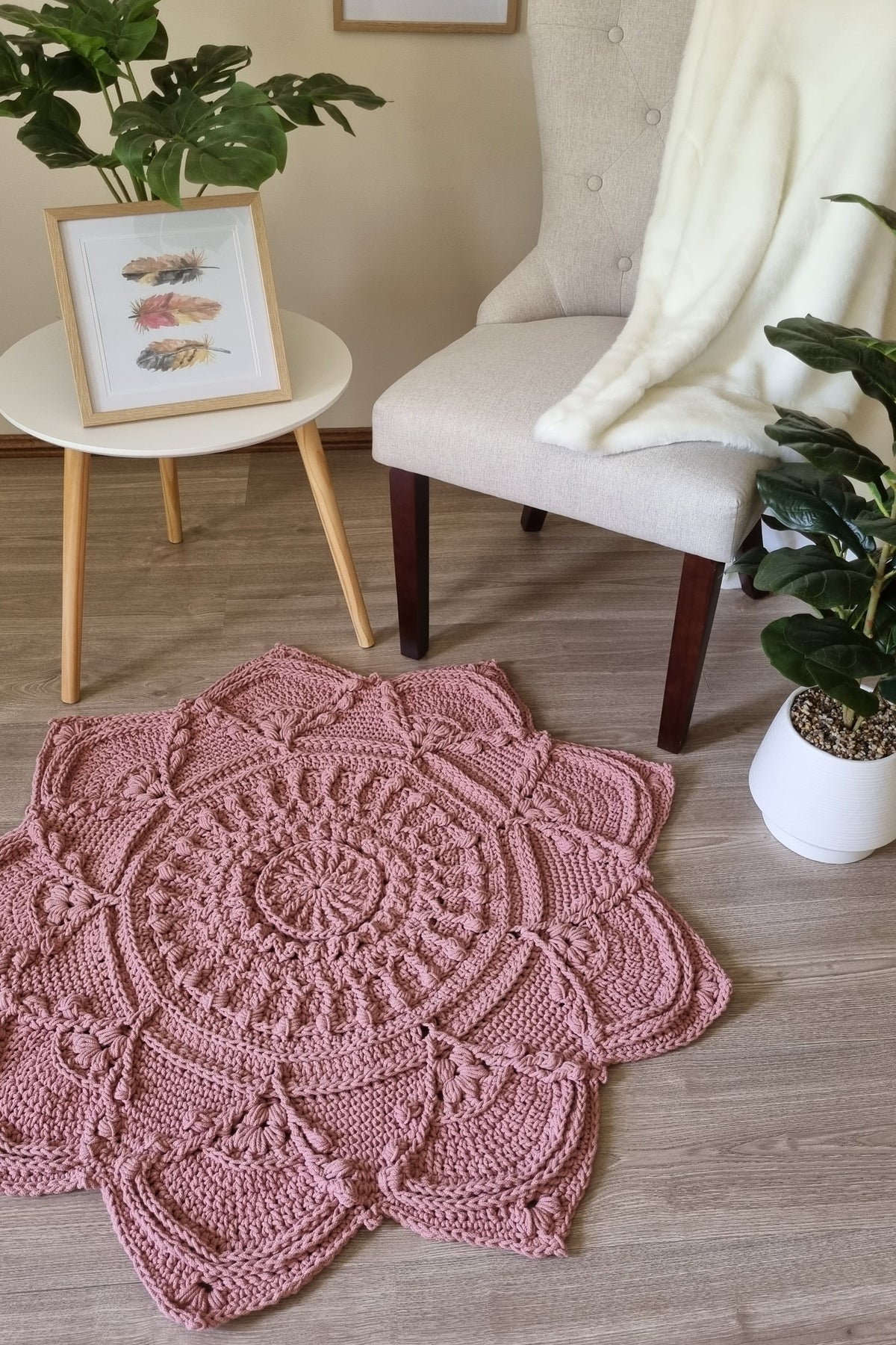 A round, hand-crocheted rug in dusty rose pink is displayed on a light wood floor in a cozy, styled room setting. The background features a beige armchair with a white throw blanket, a small white side table with a framed print and plant, and another potted plant.