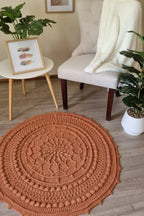 A round, hand-crocheted area rug in a warm terracotta or burnt orange color, styled in a cozy living room setting on a light wood floor next to a cream armchair and potted plants.