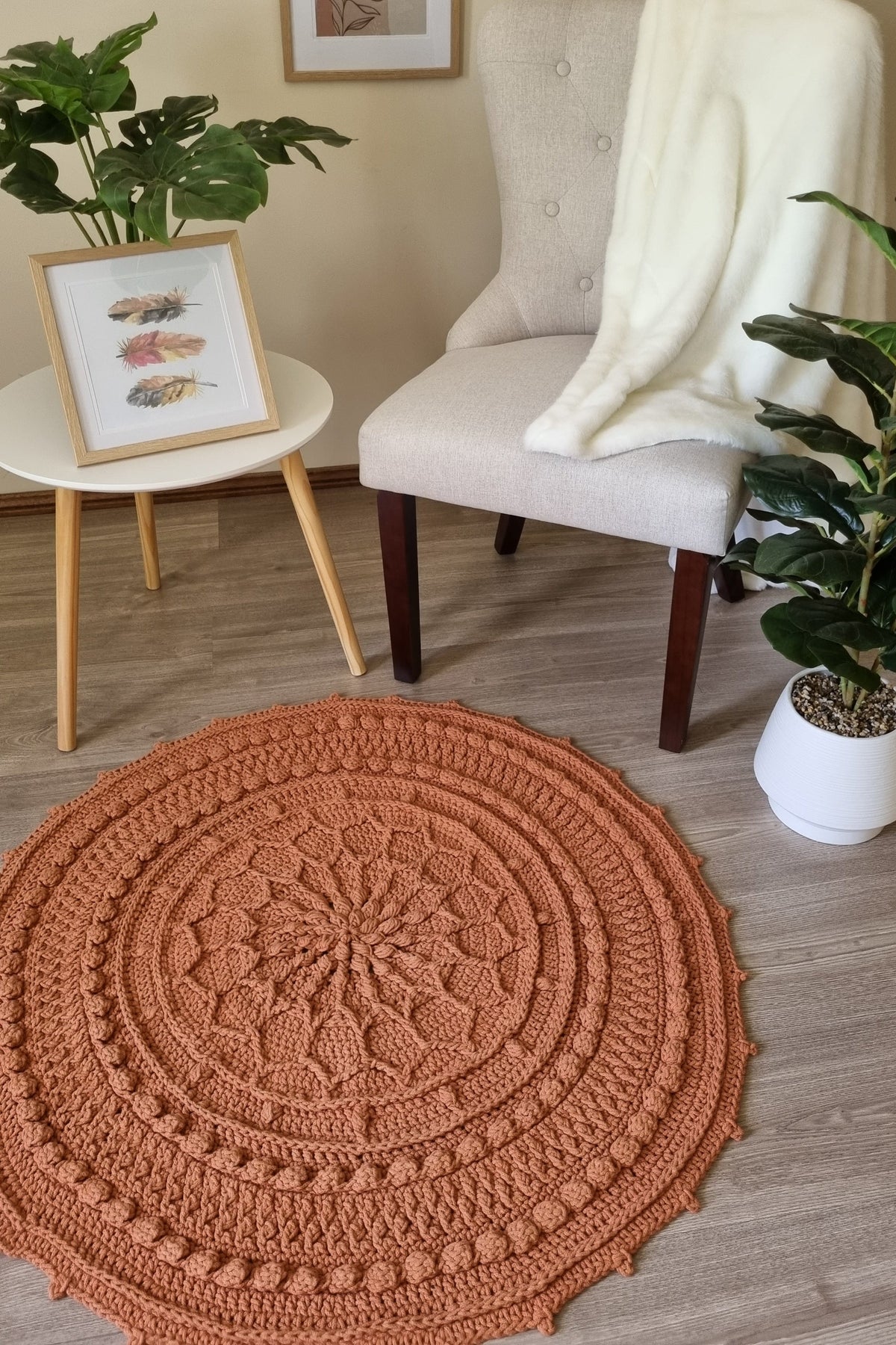 A round, hand-crocheted area rug in a warm terracotta or burnt orange color, styled in a cozy living room setting on a light wood floor next to a cream armchair and potted plants.