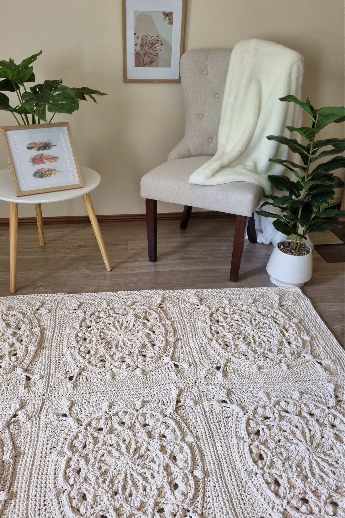 A large cream-colored crocheted area rug with intricate circular lace-like patterns, styled in a cozy living room setting with a beige armchair, side table, potted plants, and a white throw blanket.