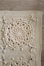 A close-up of a hand-crocheted beige floor rug laid flat on a light wood floor, showing detailed circular and floral lace patterns.