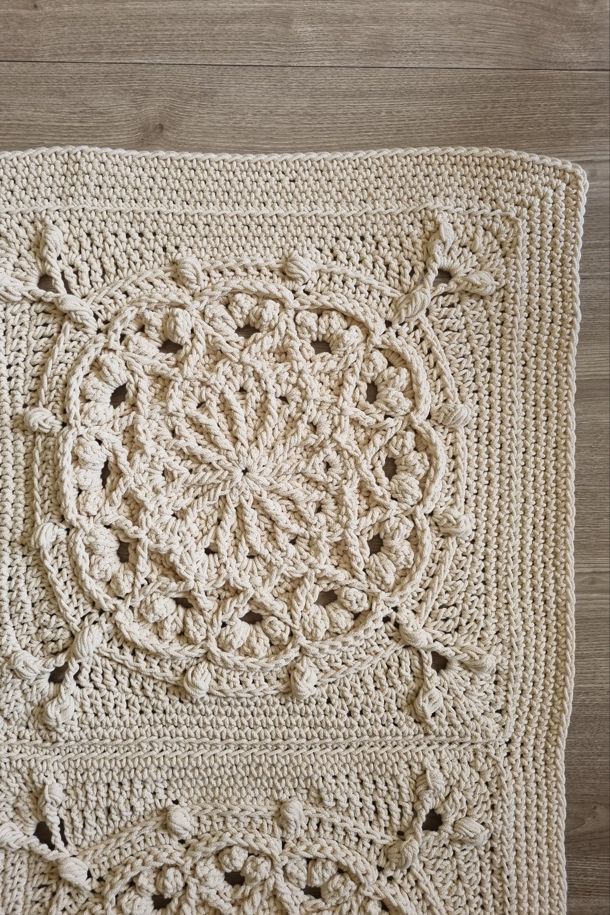 A close-up of a hand-crocheted beige floor rug laid flat on a light wood floor, showing detailed circular and floral lace patterns.