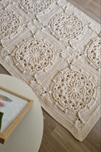 A handmade crochet area rug with a bohemian mandala pattern, laid out on a light wood floor next to a white side table with a framed print.