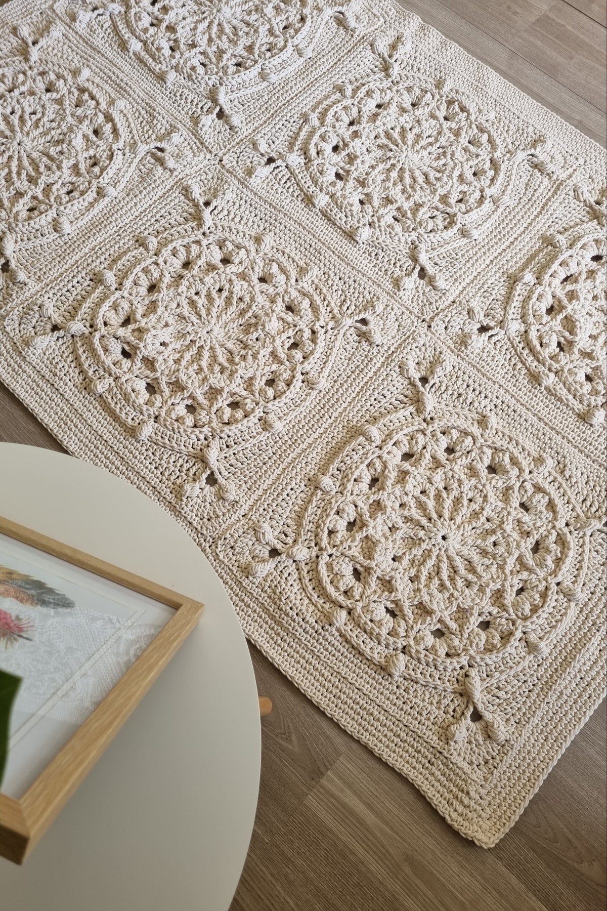 A handmade crochet area rug with a bohemian mandala pattern, laid out on a light wood floor next to a white side table with a framed print.