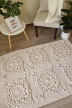 A cream-colored crocheted area rug with intricate circular lace patterns, styled in a cozy living room setting on a wooden floor next to a beige armchair and a small side table.
