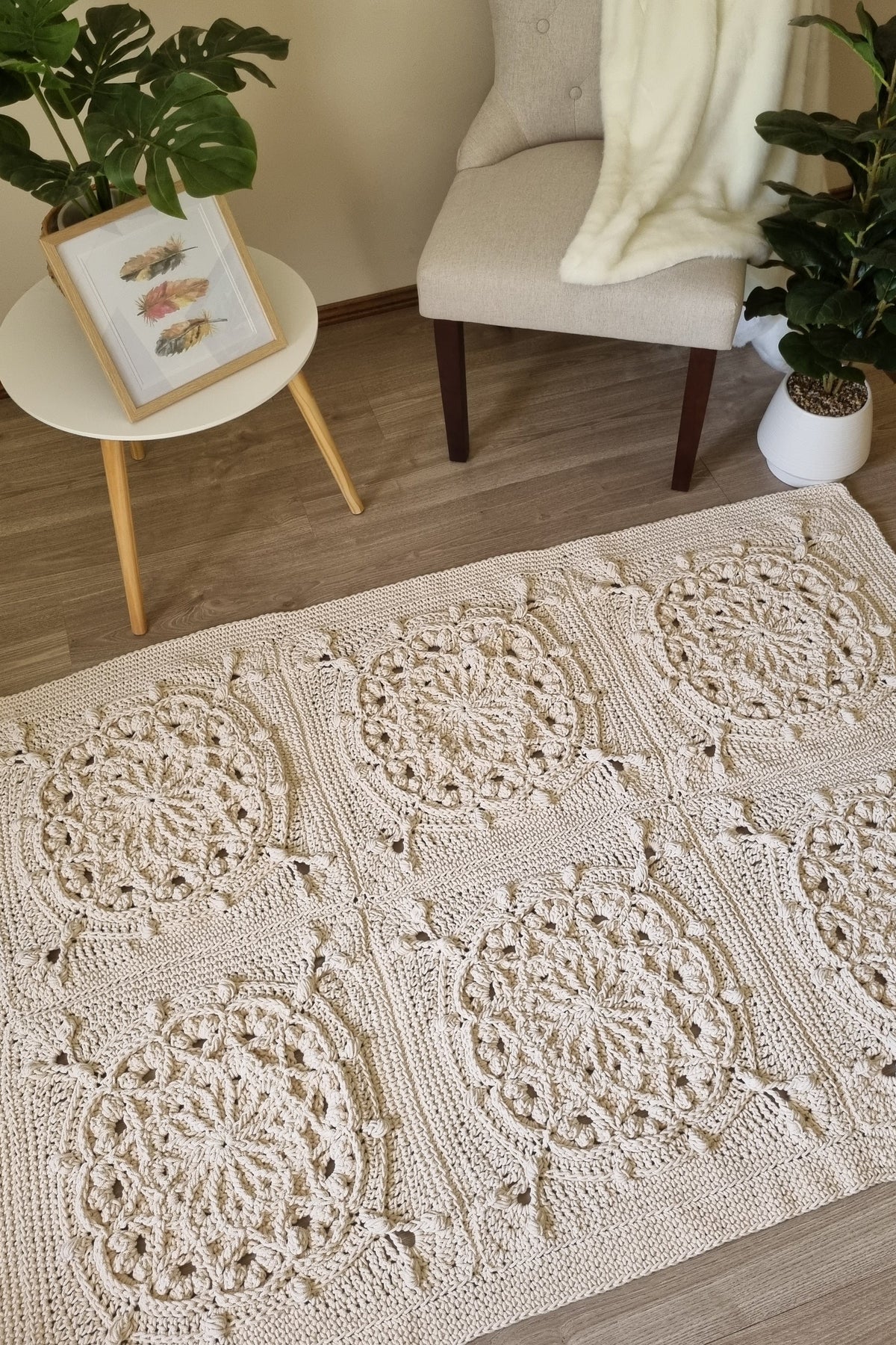 A cream-colored crocheted area rug with intricate circular lace patterns, styled in a cozy living room setting on a wooden floor next to a beige armchair and a small side table.