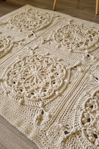 A close-up of a cream-colored crocheted area rug with intricate floral and medallion patterns, laid out on a light wood floor.