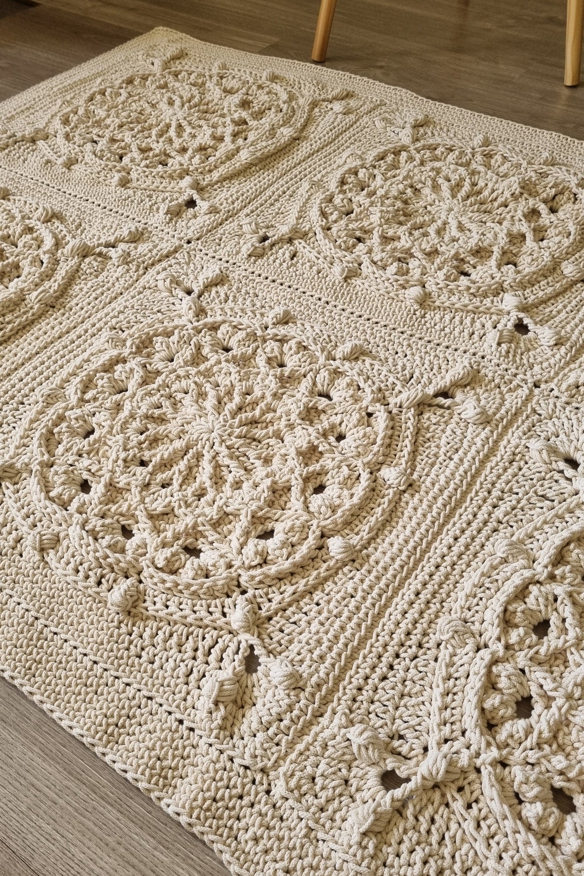 A close-up of a cream-colored crocheted area rug with intricate floral and medallion patterns, laid out on a light wood floor.