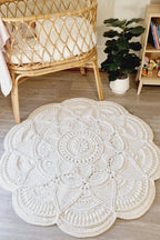 A large round crocheted area rug with an intricate mandala pattern in cream/off-white, styled on a light wood floor in a nursery setting next to a rattan bassinet and a potted plant.