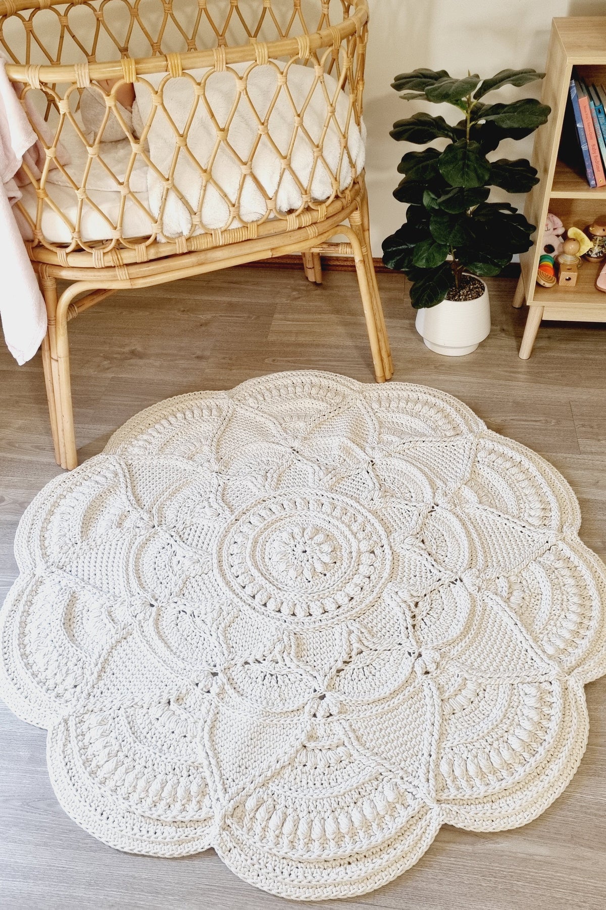 A large round crocheted area rug with an intricate mandala pattern in cream/off-white, styled on a light wood floor in a nursery setting next to a rattan bassinet and a potted plant.