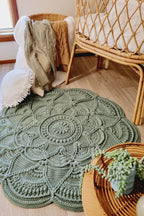 An oval, hand-crocheted sage green rug with an intricate mandala pattern, styled in a cozy nursery setting next to a rattan crib and wicker basket.
