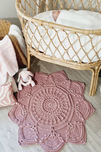 A dusty pink crocheted baby rug with a large floral or lotus-like pattern, styled on a light gray wood floor next to a rattan bassinet with a baby inside.