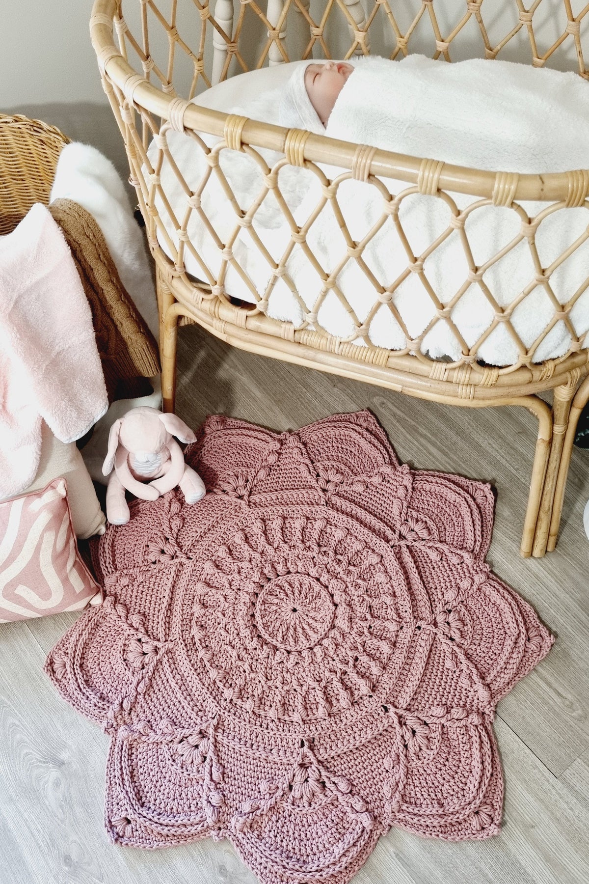 A dusty pink crocheted baby rug with a large floral or lotus-like pattern, styled on a light gray wood floor next to a rattan bassinet with a baby inside.