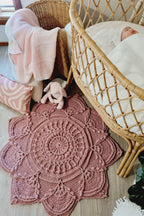 A dusty rose crocheted mandala baby rug laid out on a light wood floor next to a natural wicker bassinet with a baby inside, a wicker chair with a pink blanket, and a pink patterned pillow.