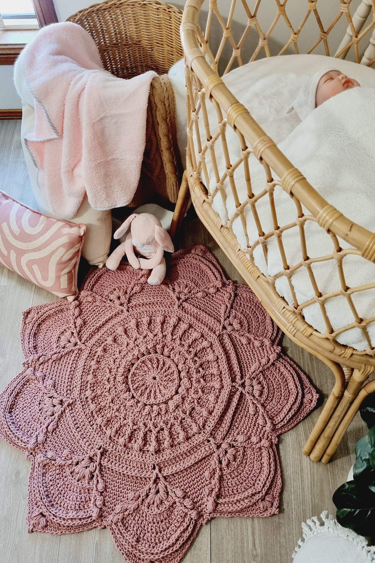 A dusty rose crocheted mandala baby rug laid out on a light wood floor next to a natural wicker bassinet with a baby inside, a wicker chair with a pink blanket, and a pink patterned pillow.