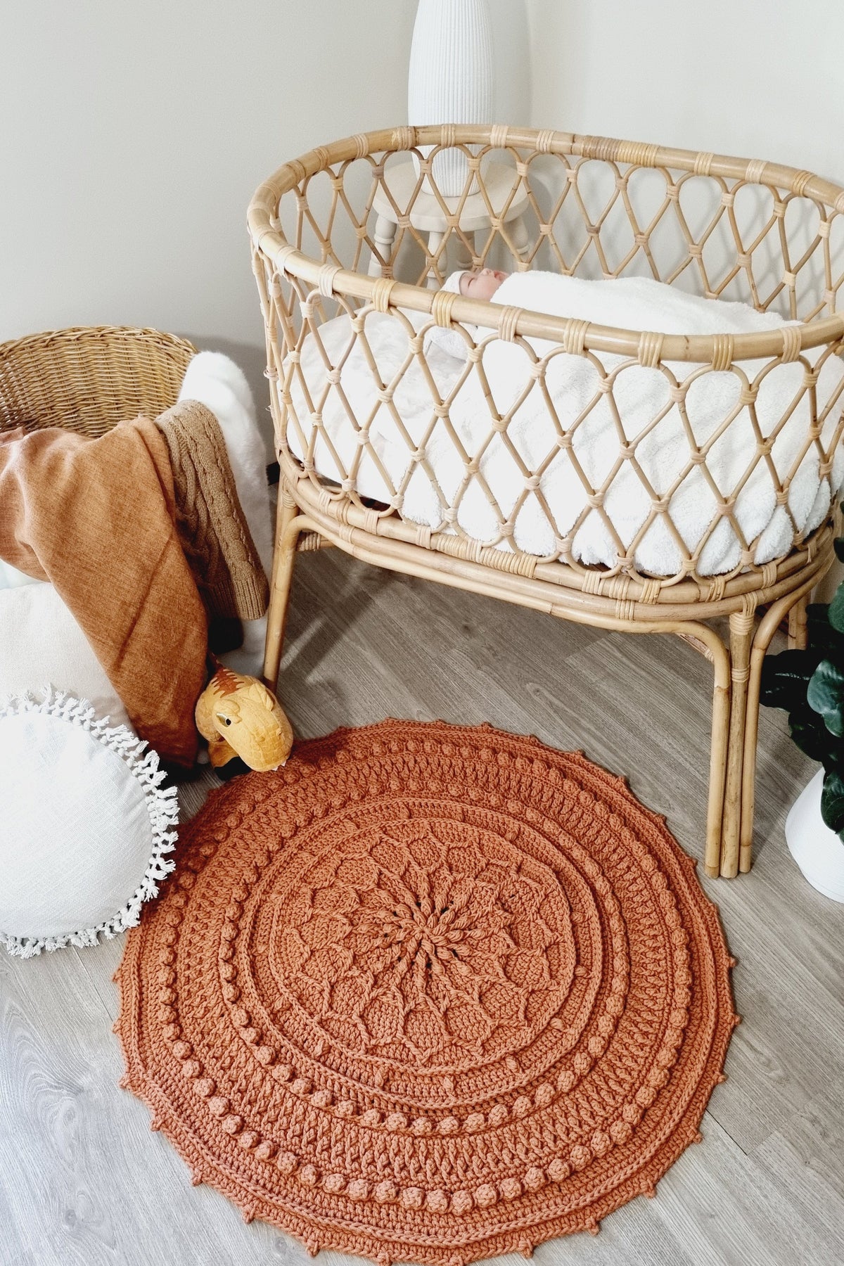 A round, rust-orange crocheted rug with a detailed mandala pattern and scalloped edge, styled on a light gray wood floor next to a rattan baby bassinet.