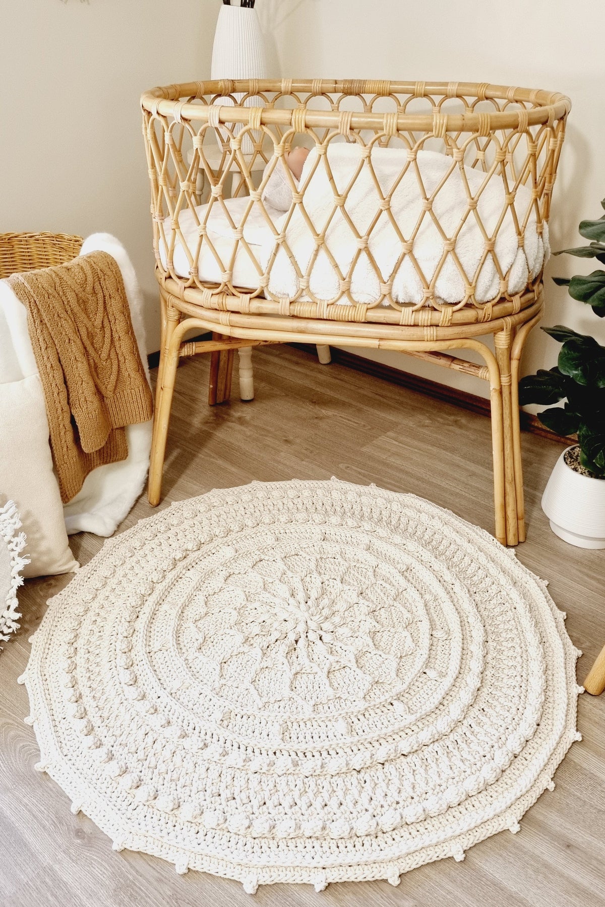 A round off-white crocheted area rug with a detailed mandala pattern and tassel trim, styled in a cozy nursery next to a natural rattan bassinet.