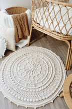 A round, cream-colored crocheted rug with a detailed mandala pattern and pom-pom trim, styled in a cozy nursery setting next to a rattan bassinet.