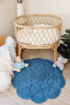 A round blue crocheted baby rug with a detailed mandala pattern, styled on a light wood floor in a nursery setting next to a rattan bassinet.