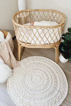 A round off-white crocheted rug with intricate mandala and floral patterns lies on a light wood floor in front of a natural rattan baby bassinet.