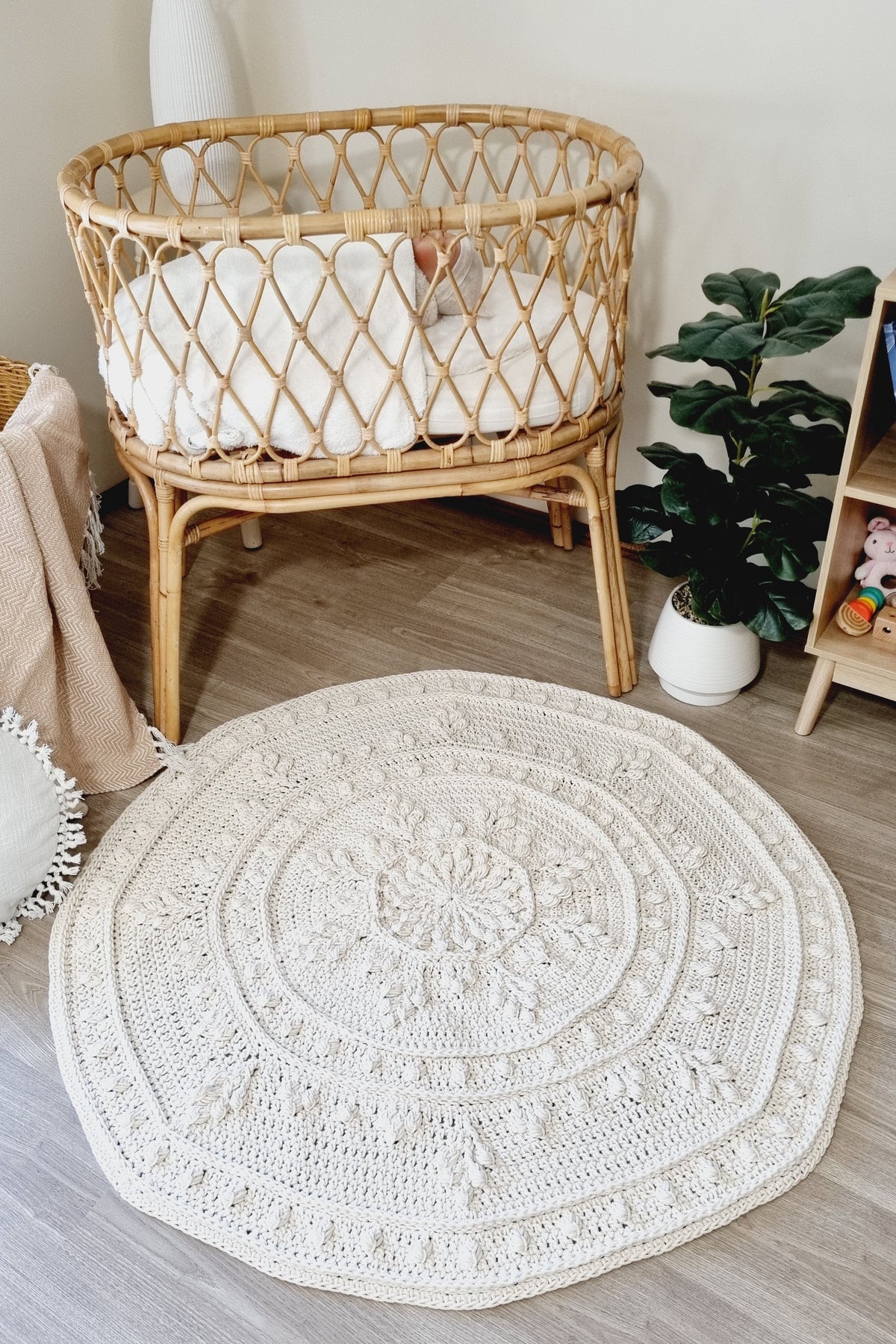 A round, off-white crocheted area rug with intricate mandala and floral patterns, styled on a light wood floor in a nursery setting next to a rattan bassinet and a potted plant.