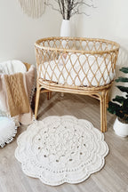 A round off-white crocheted rug with a scalloped edge and intricate floral pattern, styled in a boho nursery next to a rattan bassinet.