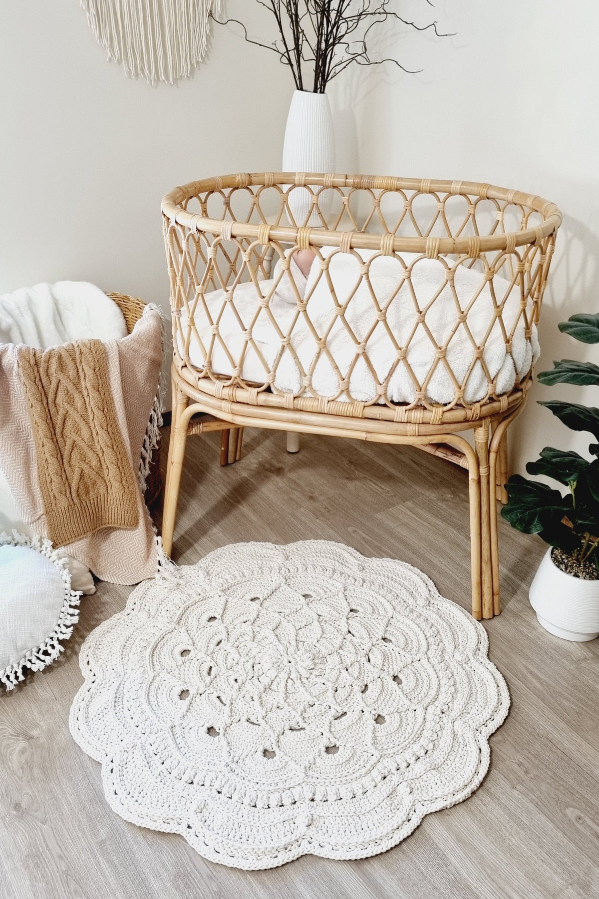 A round off-white crocheted rug with a scalloped edge and intricate floral pattern, styled in a boho nursery next to a rattan bassinet.