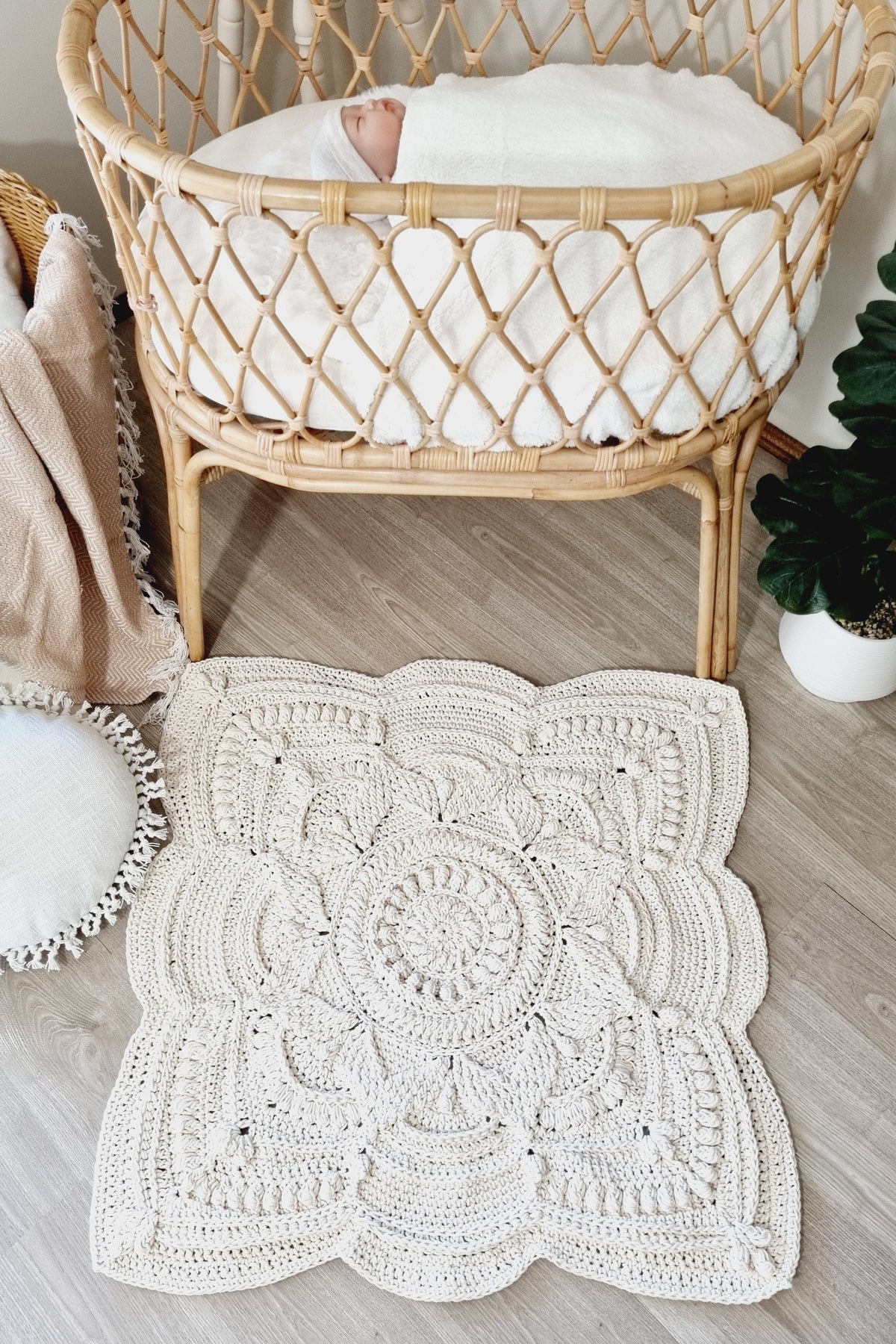 A handmade off-white crocheted baby blanket with a detailed mandala pattern and scalloped edges, styled on a light wood floor in front of a natural rattan bassinet.
