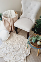 A square, cream crocheted mandala area rug styled on a light wood floor in a cozy room setting, surrounded by a tufted armchair, a basket with a beige throw blanket, and a small round side table with succulents.