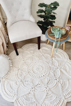 A large round off-white crocheted rug with an intricate mandala pattern, styled in a cozy room setting next to a light beige tufted armchair and a small blue side table.