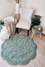 A large round crocheted rug with an intricate mandala pattern in sage green, styled on a light wood floor in a cozy room setting with a cream tufted chair, a macrame wall hanging, and a small side table.