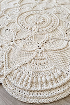 A round, intricately crocheted rug in a warm cream or off-white color, displayed on a light gray wood floor.