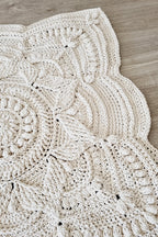 A close-up of a chunky crocheted textile with a scalloped edge, laid flat on a light wood floor. The item appears to be a cream or off-white crochet blanket, rug, or mat with textured floral and geometric patterns.