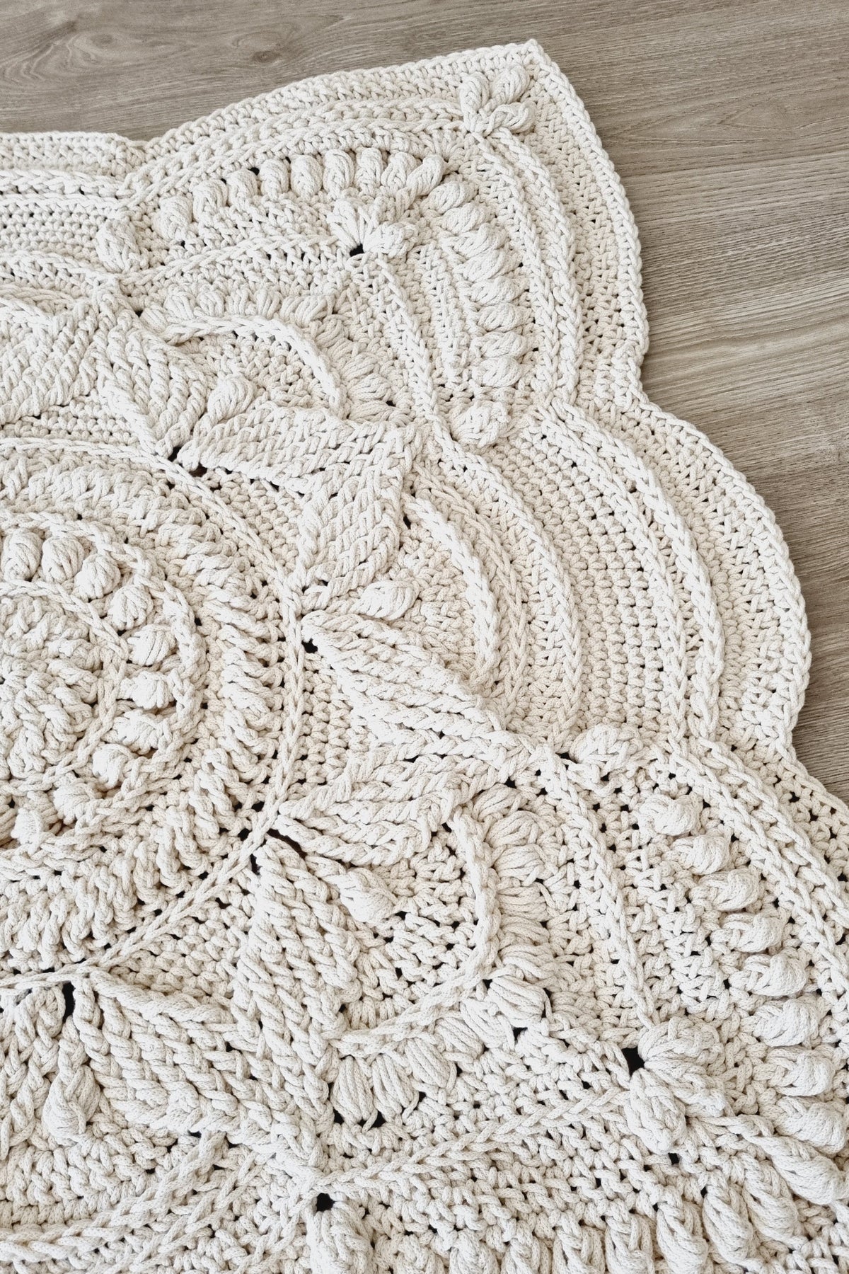 A close-up of a chunky crocheted textile with a scalloped edge, laid flat on a light wood floor. The item appears to be a cream or off-white crochet blanket, rug, or mat with textured floral and geometric patterns.