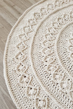 A close-up of a round, hand-crocheted rug in a warm cream or off-white color, laid out on a light wood floor. The rug features a detailed, textured cable-knit pattern with concentric circular and diamond-like motifs.