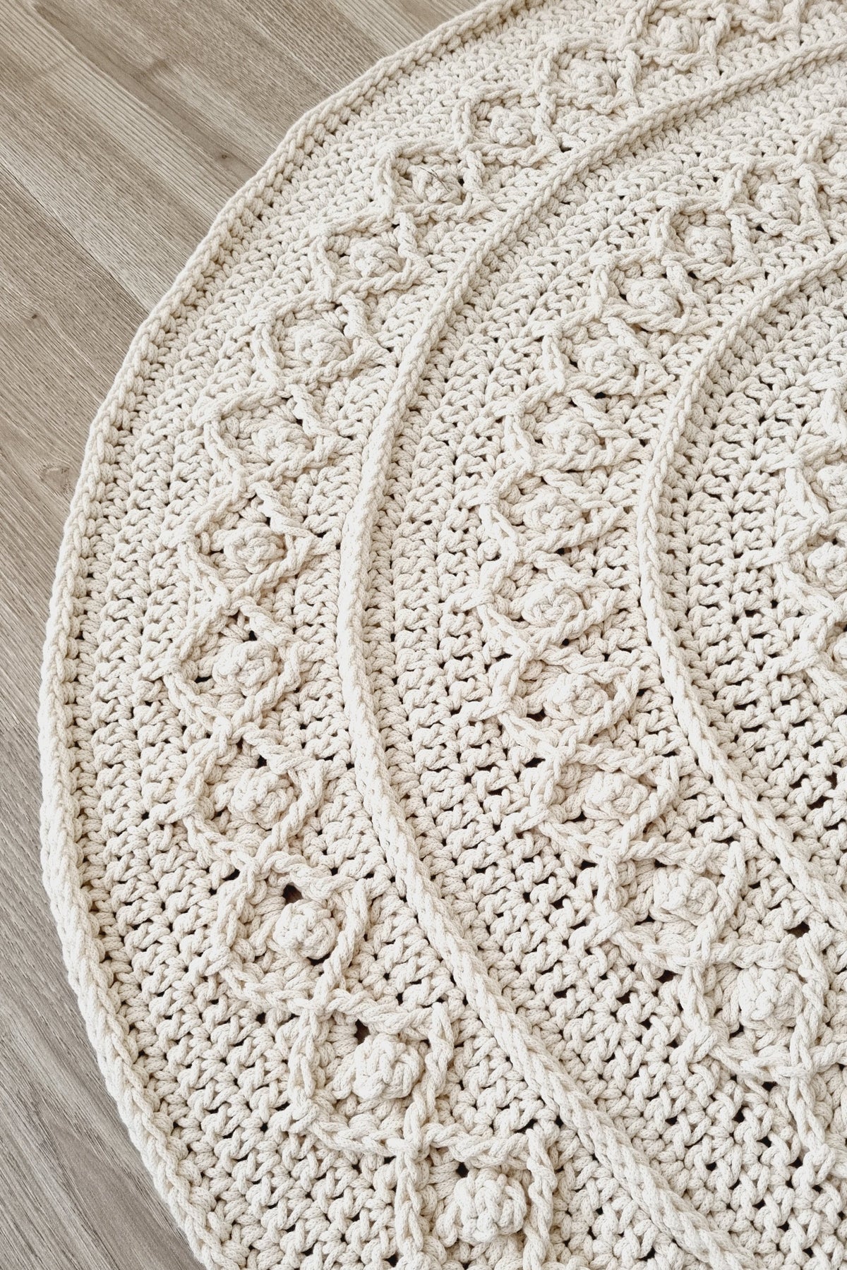 A close-up of a round, hand-crocheted rug in a warm cream or off-white color, laid out on a light wood floor. The rug features a detailed, textured cable-knit pattern with concentric circular and diamond-like motifs.