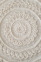 A close-up of a round crocheted textile with concentric mandala-style patterns in a warm off-white or cream tone.