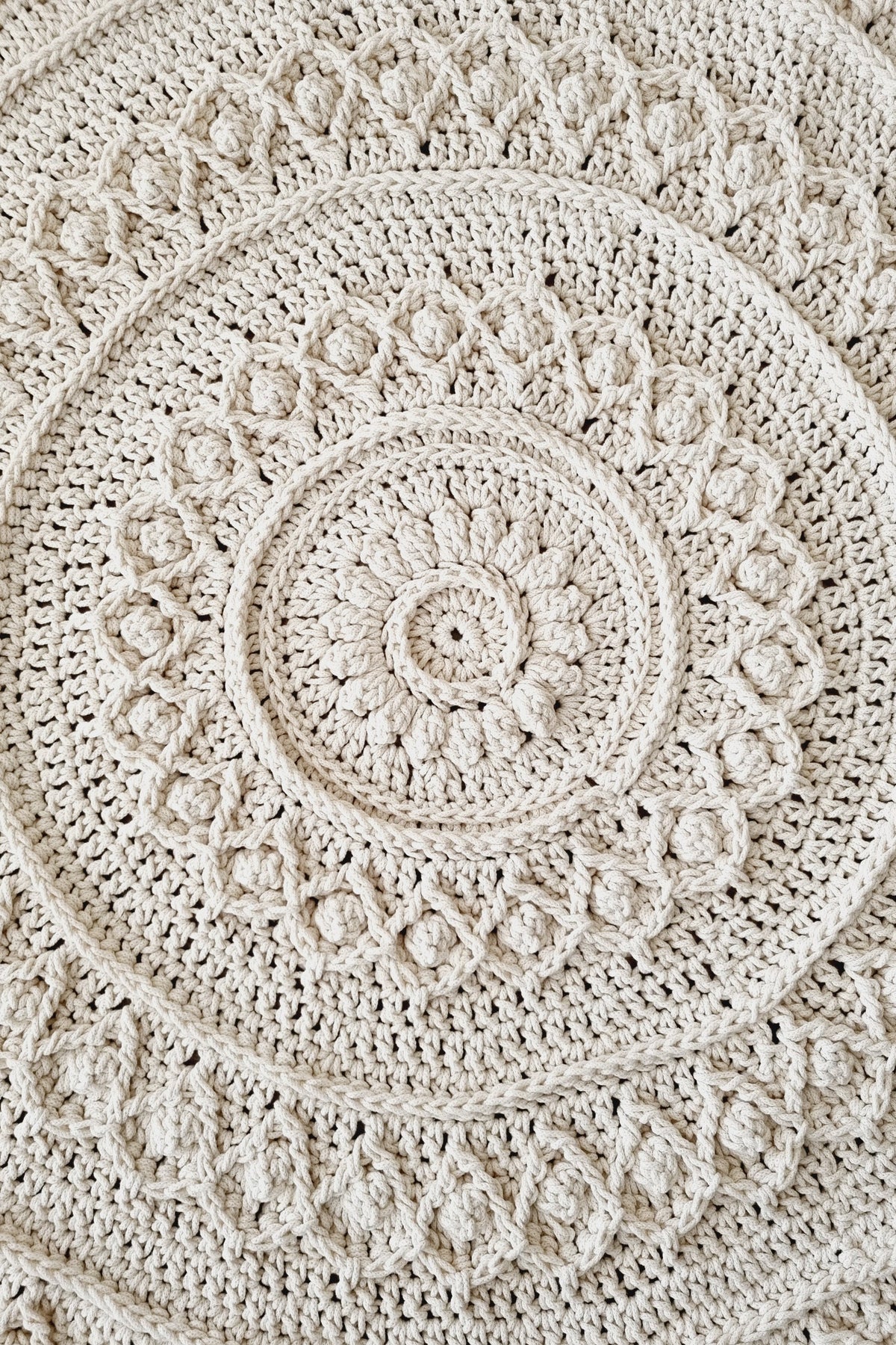 A close-up of a round crocheted textile with concentric mandala-style patterns in a warm off-white or cream tone.